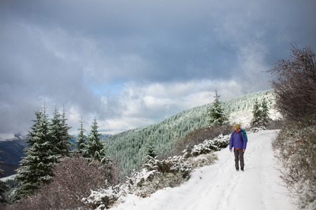 a woman walks and leaves footprints in the snow, young woman with a backpack walking along the trail in the mountains on the background of snowy mountains and cloudy sky.の写真素材
