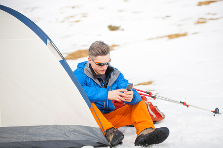 climber with glasses sits in a tent near a backpack in the winter, man holding mobile phone, the tent stands in the snow.の写真素材
