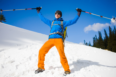 a young man with trekking poles and backpack goes on the snow in the mountains against the blue sky and clouds, mountaineer in a blue jacket and orange pants goes over the white snow in the mountains.の写真素材