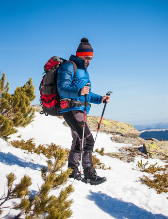 Man with trekking poles goes up in the mountains in winter, the climber with a backpack walking on snow on a Sunny day on the background of blue sky.の写真素材