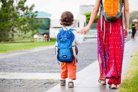 A child with a backpack walks with mother's hand. A curly-haired boy walks through the park and holds mother's hand. Mom and son are walking along the road.の写真素材