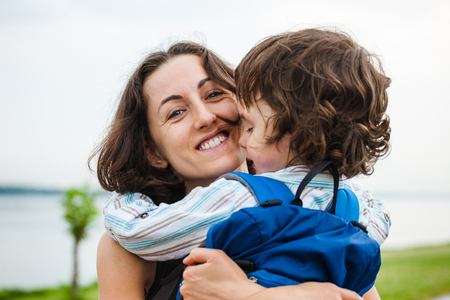 A woman hugs a curly-haired boy and smiles. Mom feels sorry for her son. Meeting mom and baby after separation. A child with a backpack hugs her mother.の写真素材