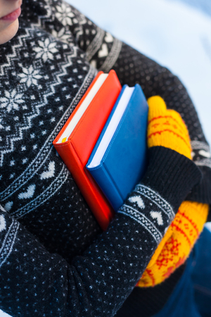 The girl in the knitted sweater and mittens holds two books against the winter snow.の写真素材