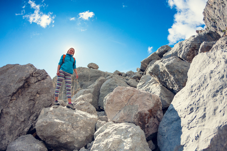 A girl with a backpack walks along the mountain path against a beautiful blue sky with clouds. A woman walks along a stony path. A blonde is traveling with a backpack.の写真素材