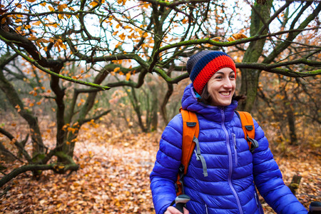 A girl with trekking sticks walks along a forest trail. Skyrunning in the fall. Active recreation in nature. A woman is walking on a dirt road. Portrait of a smiling girl in a knitted cap.の写真素材