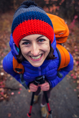 A girl with a backpack walks along a forest trail. Skyrunning in the fall. Active recreation in nature. A woman is walking on a dirt road. Portrait of a smiling girl in a knitted cap.の写真素材