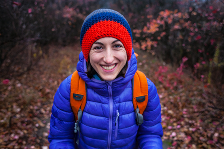 A girl with a backpack walks along a forest trail. Skyrunning in the fall. Active recreation in nature. A woman is walking on a dirt road. Portrait of a smiling girl in a knitted cap.の写真素材