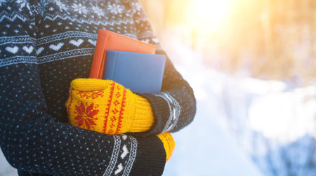 The girl in the knitted sweater and mittens holds two books against the winter snow.の写真素材
