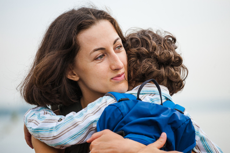 A woman hugs a curly-haired boy and smiles. Mom feels sorry for her son. Meeting mom and baby after separation. A child with a backpack hugs her mother.の写真素材