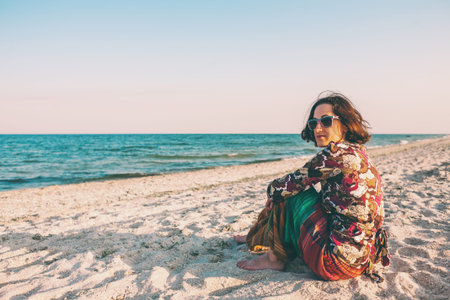 A girl in Indian clothes on the sea coast. A woman sits on the sand near the ocean. A woman sits alone at sunset and admires the surf.の写真素材