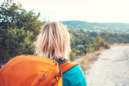 A girl with a backpack walks along the mountain path. A woman is walking along a stony road. Blonde with an orange backpack.の写真素材