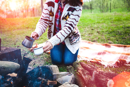 A girl is drinking coffee by the bonfire. A woman is preparing a morning coffee on fire. The brunette pours the drink into the cup. Camping in the forest. Picnic in nature.の写真素材