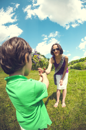 The boy gives his mother a bouquet of wild flowers. A woman is walking with her son in a meadow. A gift for mother's day. The girl takes a bunch of flowers. The child congratulates mom.の写真素材