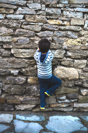 A restless child. Fidget. The boy climbs the ancient stone fence. The child is trying to climb a stone wall. An old brick wall. A little rock climber. Childish curiosity.の写真素材