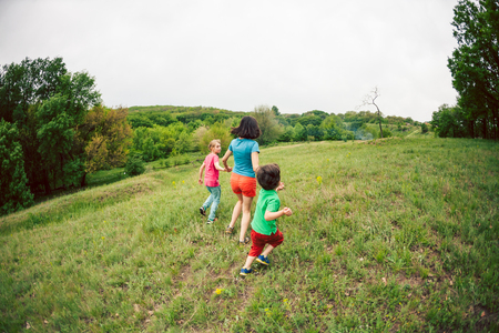 A woman with children runs along the green grass. Mom holds her sons by the hands and run. A boy playing with his family outdoors. The kid walks through the meadow with his mother and brother.の写真素材