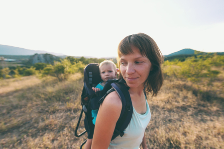 A boy is sitting in a backpack. The kid is traveling with his parents. Climbing with a child. The mother carries the baby. A little traveler. A woman is carrying a backpack with her baby.の写真素材