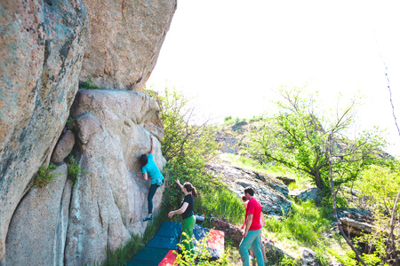 Climbing in nature. Friends climb to the stone. The girl climbs on the stone, and friends support her. Bouldering in nature. Active lifestyle. Sports people. Gymnastic insurance.の写真素材