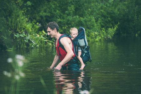 A man with a child crosses the river. The father carries the son in a backpack. Rescue from flooding. Evacuation of flood victims in Asia. Traveling with a child through the jungle. Extreme tourism.の写真素材