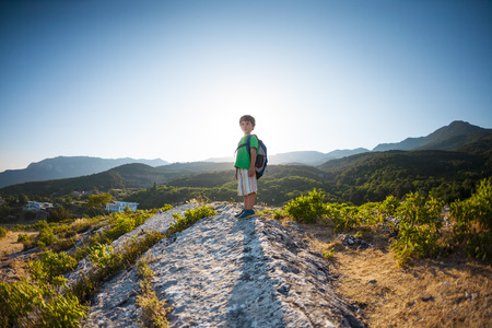 Boy at the top of the mountain at sunset. A child with a backpack travels to interesting places. The kid walks along the mountain path. Active vacations. A little traveler. Climbing.の写真素材