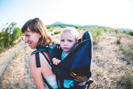 A boy is sitting in a backpack. The kid is traveling with his parents. Climbing with a child. The mother carries the baby. A little traveler. A woman is carrying her baby. Traveling with children.の写真素材
