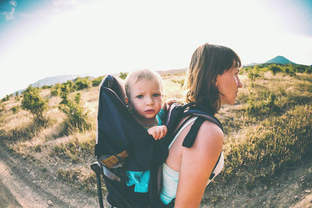 A boy is sitting in a backpack. The kid is traveling with his parents. Climbing with a child. The mother carries the baby. A little traveler. A woman is carrying a backpack with her baby.の写真素材