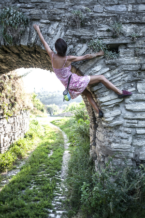 The girl climbs the stone wall. A woman in a summer dress climbs the wall of an old destroyed building. Brick fence. The climber is hanging on the city building. Strengthening the ruined castle.の写真素材