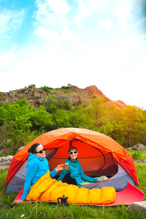 Two girls are sitting in a tent. Girlfriends communicate in nature. Women in sleeping bags drink coffee. Friends in the campsite. Smiling brunettes talking.の写真素材