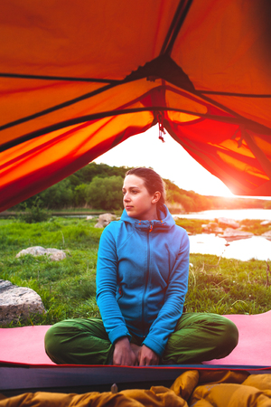 The girl is sitting near the tent. A woman in a sleeping bag. A smiling brunette is resting in nature. Camping by the river.の写真素材