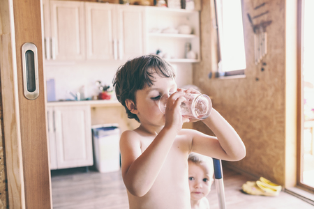 The boy drinks water from a glass. Children quench their thirst on a hot summer day. The brothers play together at home. Naked kids run around the house. The child is cleaning in the kitchen.の写真素材