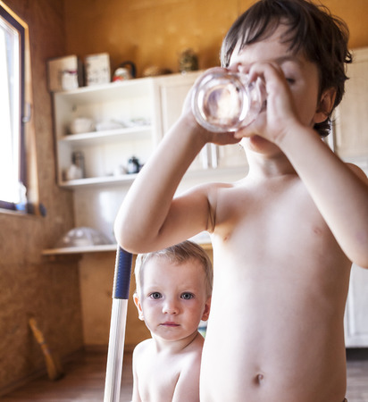 The boy drinks water from a glass. Children quench their thirst on a hot summer day. The brothers play together at home. Naked kids run around the house. The child is cleaning in the kitchen.の写真素材