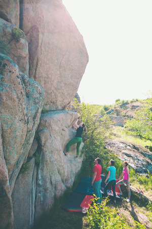 Climbing in nature. Friends climb to the stone. The girl climbs on the stone, and friends support her. Bouldering in nature. Active lifestyle. Sports people. Gymnastic insurance.の写真素材