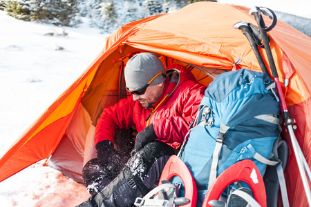 Camping in the snow. Winter trekking in the mountains. Orange tent, snowshoes and trekking poles. A man with a backpack sets a tent on the snow. Extreme journey. Equipment for winter hiking.の写真素材