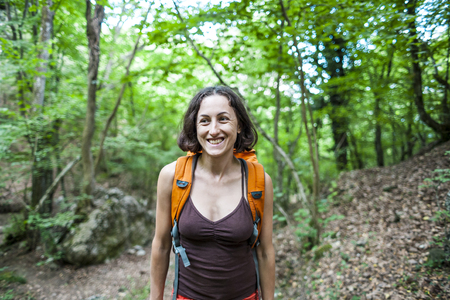 Girl with a backpack in the forest. A woman is walking along a mountain path. Travel alone. Smiling brunette against a background of green trees.の写真素材