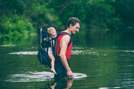 A man with a child crosses the river. The father carries the son in a backpack. Rescue from flooding. Evacuation of flood victims in Asia. Traveling with a child through the jungle. Extreme tourism.の写真素材