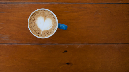 Coffee mug on a wooden table. Heart shape pattern on coffee creme. Morning refreshing drink in a blue cup.の写真素材