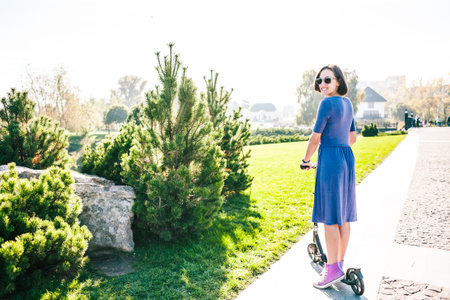 Woman riding a scooter. The girl in a dress rides a scooter in the park. Brunette pushes off the...