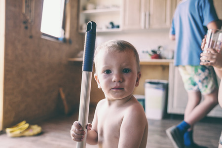 A little boy helps parents clean up in the kitchen. The kid is holding a mop. The child washes the floor with a mop. Naked baby is learning to help his mother.の写真素材