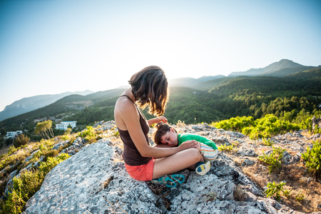 The tired boy lies on his mother's feet. A woman is traveling with her son. Tiring climb to the top of the mountain. Mom hugs the child, sitting on top of the mountain. Baby is sleeping.の写真素材