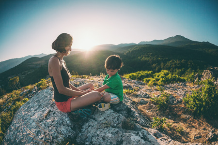 A woman is traveling with a child. Mother with a son is sitting on a large rock. Climb to the top of the mountain with children. The boy with the backpack climbed to the top. Active vacations.の写真素材
