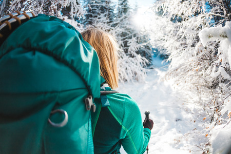 Woman with backpack in the winter mountains. Travel to scenic places. Portrait of a blonde with trekking sticks. A tourist walks through a snowy forest. Active leisure and lifestyle. Smiling girl.の写真素材