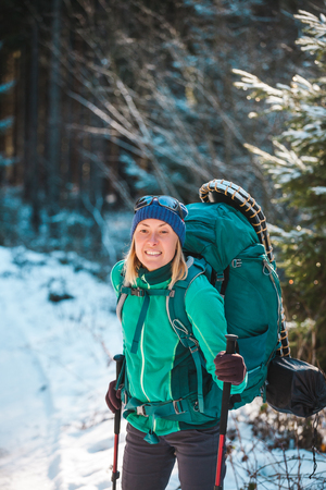 Woman with backpack and snowshoes in the winter mountains. Travel to scenic places. Portrait of a blonde with trekking sticks. A tourist walks through a snowy forest. Girl walks along the path.の写真素材