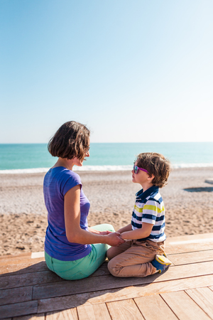 Woman playing with her son on the beach. The boy with his mother sitting on the ocean shore. A child hugs his mother. Communication mother and child.の写真素材