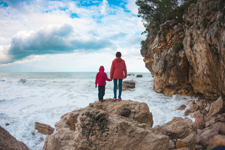 A woman with her son are standing on a stone and looking at the sea. The boy with his mother look at the waves. The child is walking along the ocean shore. Silhouette of mom and child against the sky.の写真素材