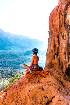 Climber sits near the rope and prepares to overcome the route. Slender girl holds climbing equipment. A woman in a helmet on the background of beautiful mountains. Rest after climbing.の写真素材