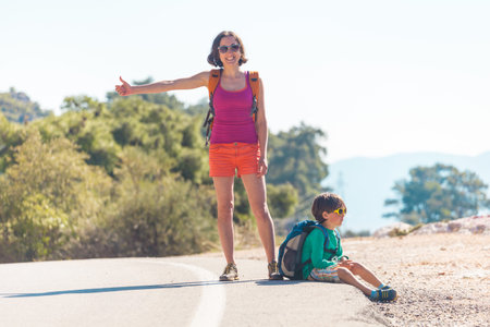 Woman with a child hitchhiking. The girl catches the car. Brunette with a backpack and young son waiting for transport. Hitchhiking in Turkey. The boy with his mother stands on a mountain road.の写真素材