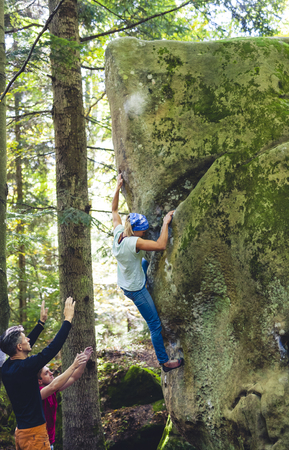 Bouldering in nature. Climbing on natural terrain. Safety in rock climbing. Friends train in nature. Active lifestyle. Teamwork. A woman climbs on a big stone, and friends insure her.の写真素材