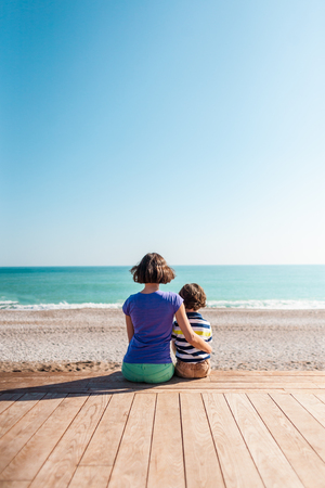 Woman playing with her son on the beach. The boy with his mother sitting on the ocean shore. A child hugs his mother. Communication mother and child.の写真素材