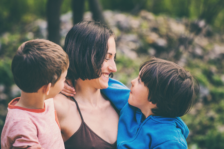 Woman hugging sons on the terrace of a wooden house. Boys spend time with mom. Young mother walks with children. A happy family. Young brunette fooling around with a child.の写真素材