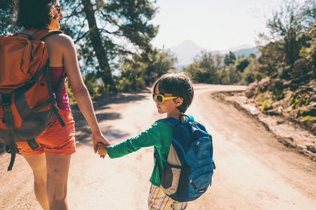 The boy walks with his mother on a mountain road. A woman travels with a child. The kid holds mom's hand. Travel with backpacks. Hike and climb with kids.の写真素材
