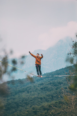 A man is walking along a stretched sling. Highline in the mountains. Man catches balance. Performance of a tightrope walker in nature. Highliner on the background of valley.の写真素材